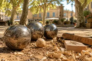 Polished pétanque balls and wooden accessories on sunlit earth with wood shavings and a cochonnet, evoking Provençal warmth and artisanal detail.